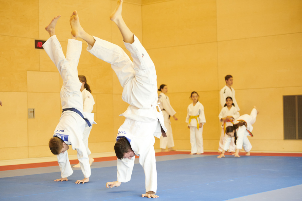Zwei jugendliche Judoka machen beim Aufwärmen einen Handstand, im Hintergrund dehnen und warten weitere Kinder auf der Matte