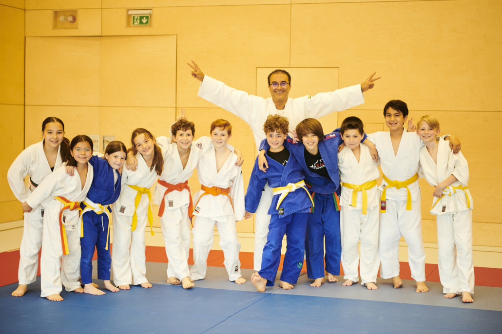 Kinder in weißen und blauen Judoanzügen jubeln mit erhobenen Armen, in der Mitte ihr Trainer Afshin Karimian